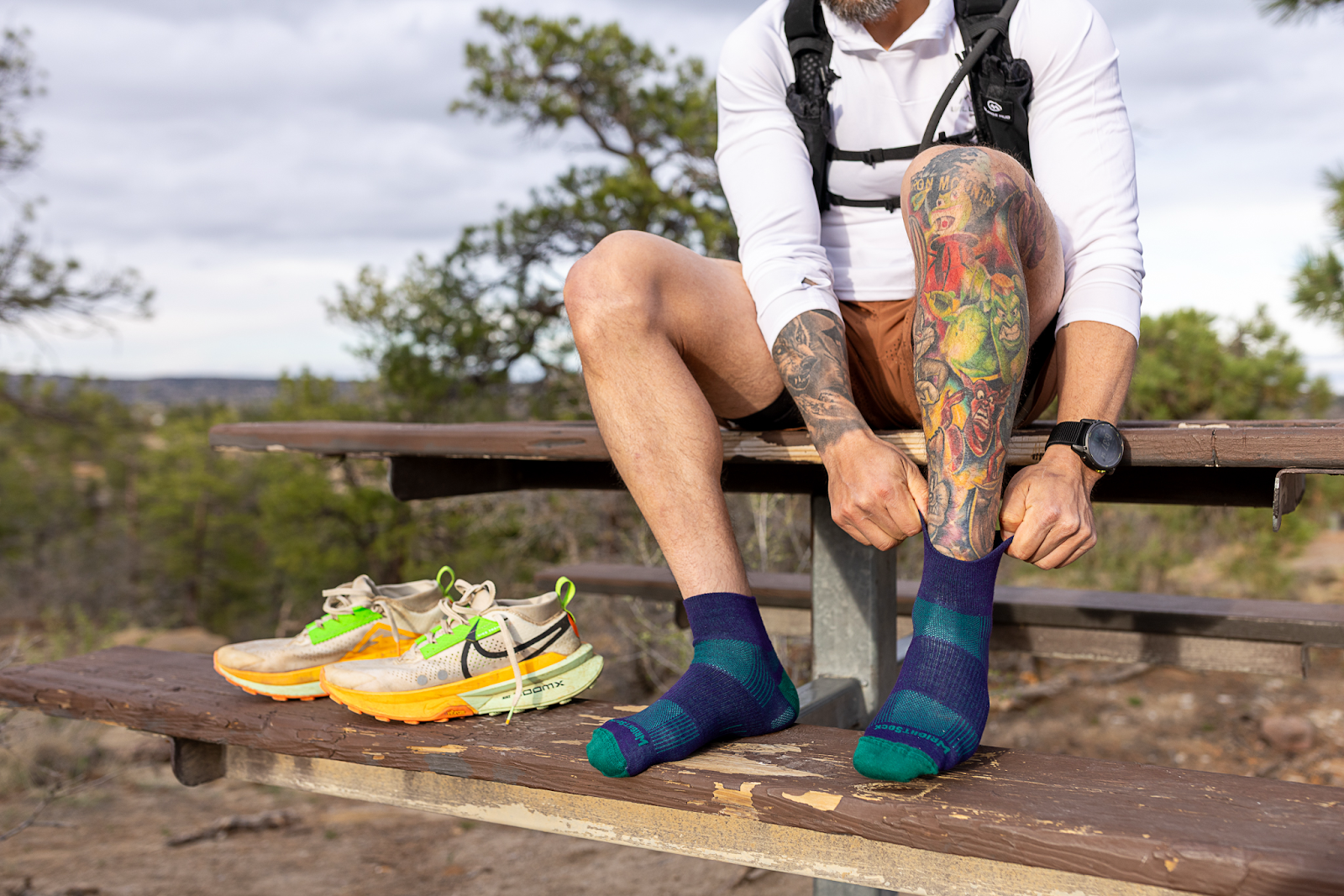 hiker sitting on wooden bench putting on blue striped crew socks with running shoes beside him