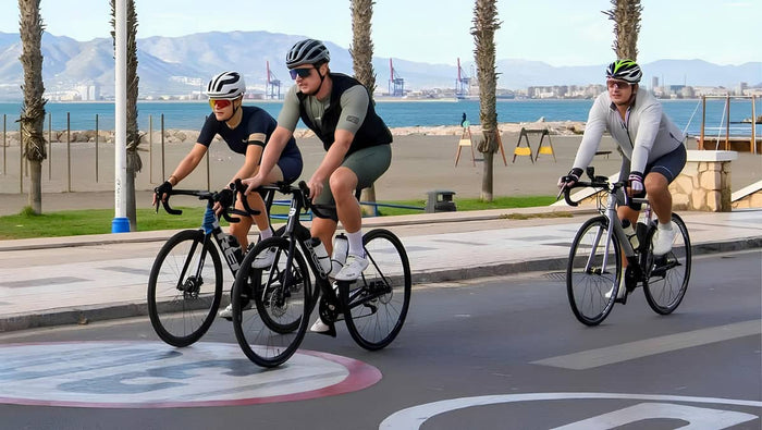 Three cyclists riding on a road near a waterfront with palm trees and mountains in the background.